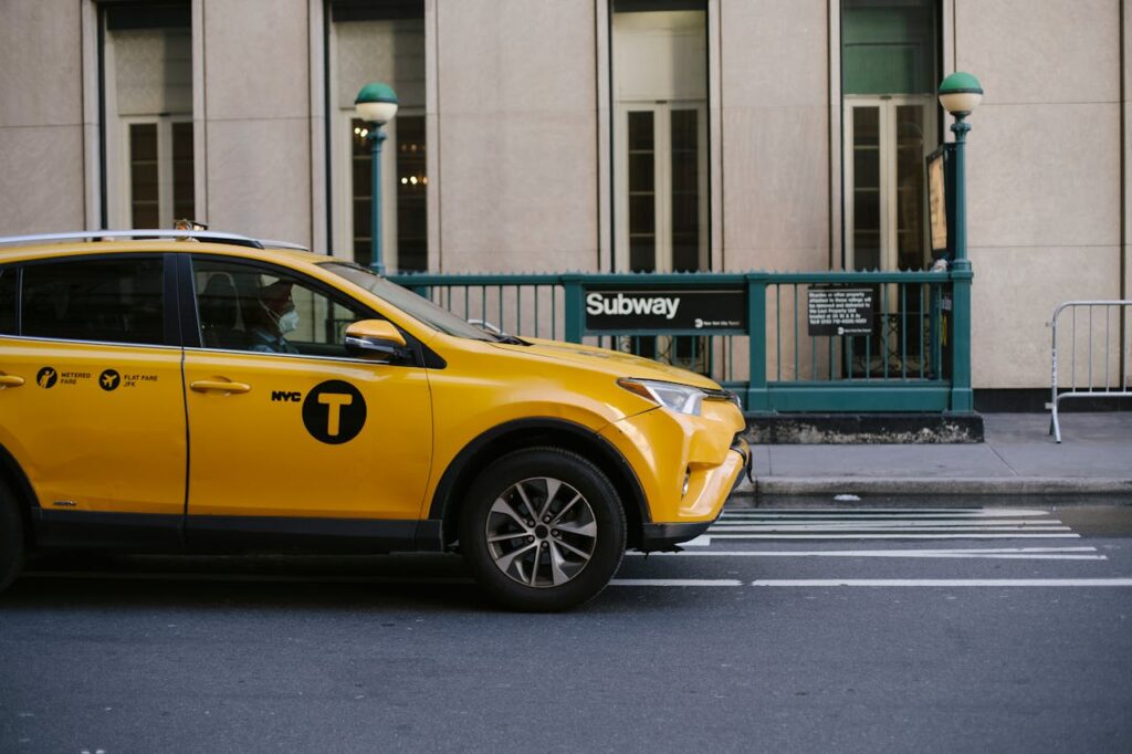 Contemporary yellow taxi with driver in medical mask standing on empty road near modern building and subway station entrance during coronavirus pandemic in New York USA